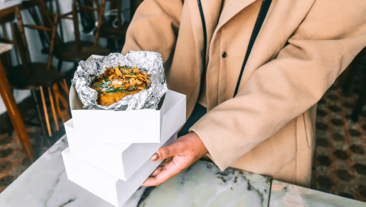 Person holding a takeout box with food wrapped in foil on top.