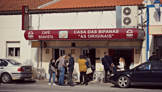 People gather outside a café with red awning on a sunny day.