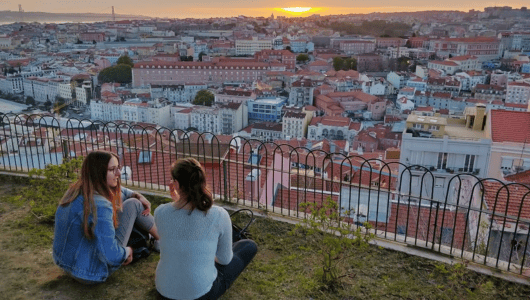 Two people sitting on grass, overlooking a cityscape at sunset.