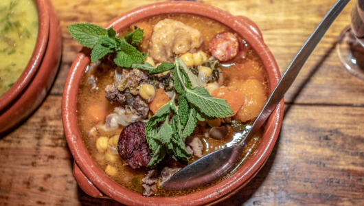 Clay bowl with hearty stew, including meat, corn, and greens, on wooden table.