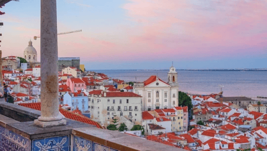 Scenic view of city with red rooftops, sea, and pink sky at sunset.