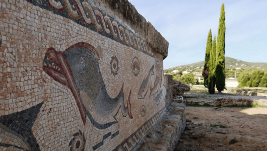 Ancient mosaic wall with dolphin design, next to a cypress tree.