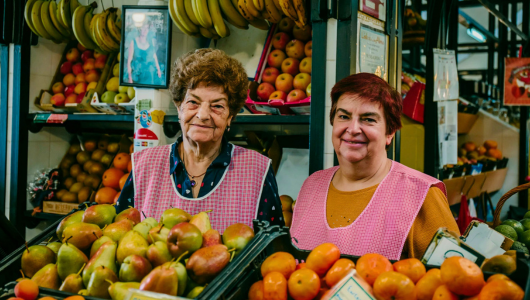 Two women in pink aprons at fruit market stall with pears and oranges.