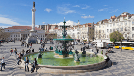 People around a fountain in a European square with colorful buildings.