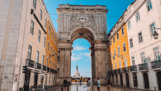 People walking toward a large archway on a sunny street between two colorful buildings.