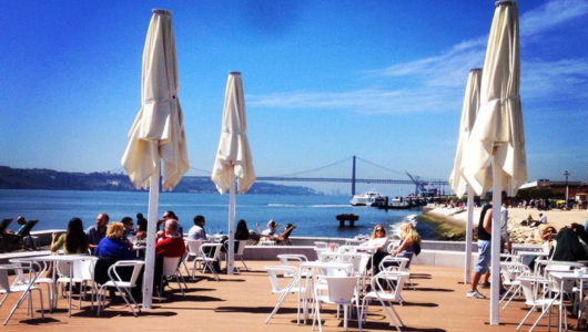 Outdoor cafe with white umbrellas near a river and bridge views.