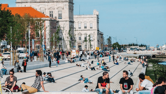 People sitting on a wide riverside promenade with historic buildings nearby.