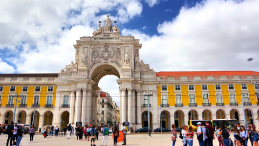 People in a plaza in front of a large ornate archway under a cloudy sky.