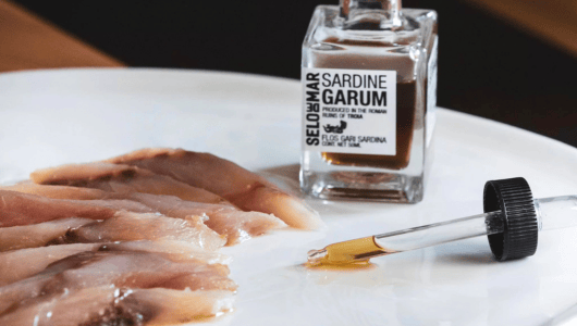 Sliced fish with a dropper and jar of sardine garum on a white plate.