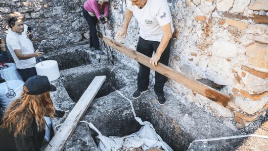Archaeologists study excavation site with stone walls and wooden beams.