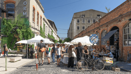 People gather at an outdoor market with umbrellas and brick buildings.