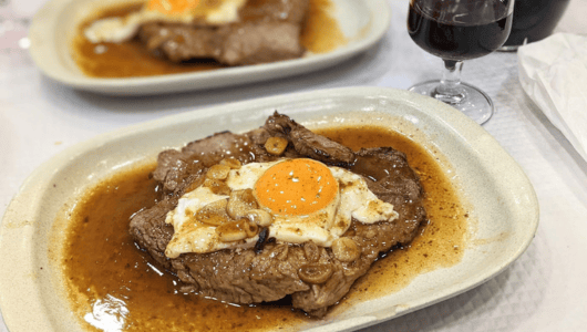 Steak with fried egg and sauce served on a white plate, glass of red wine nearby.